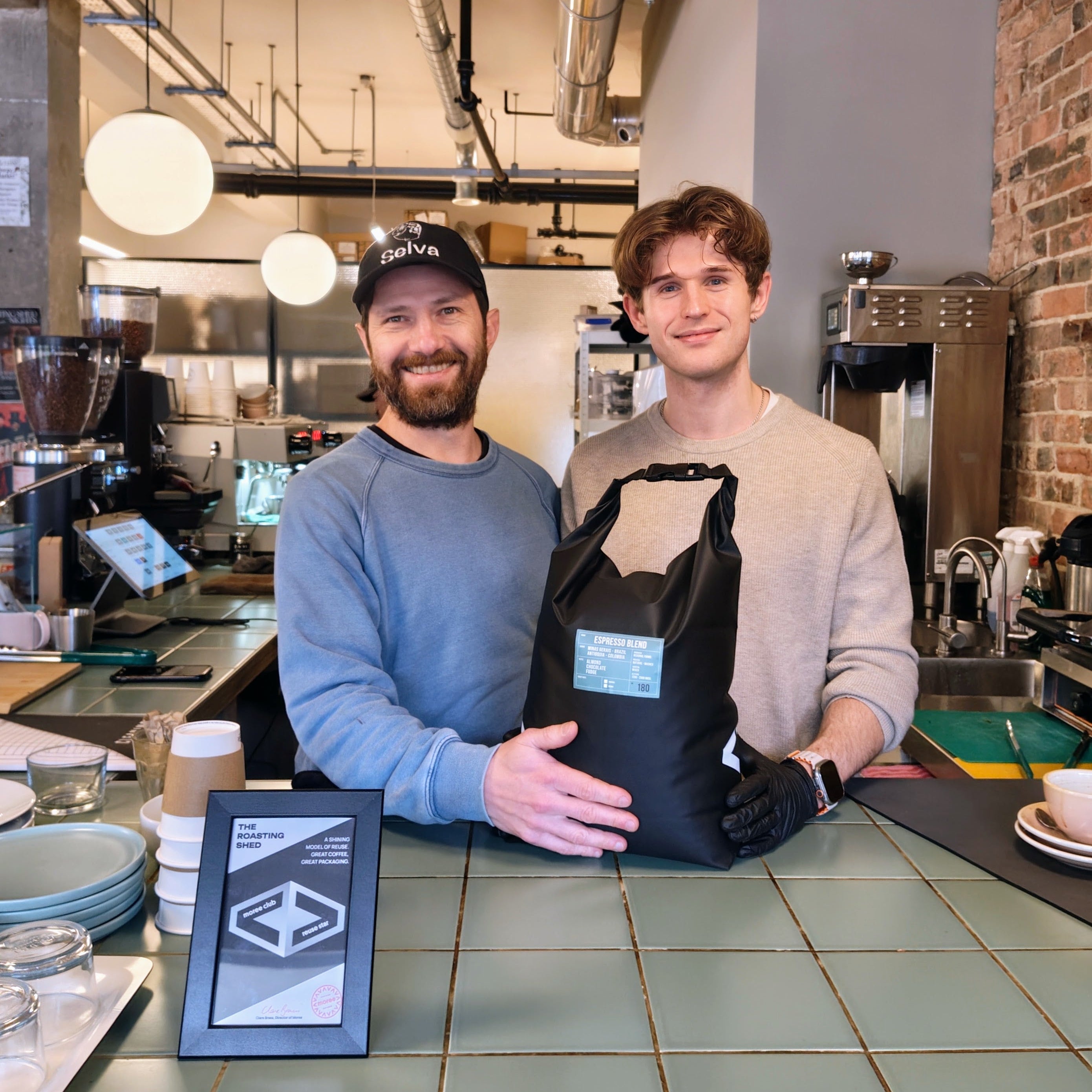 Barista and roaster holding a black reusable moree bag in a coffee shop setting